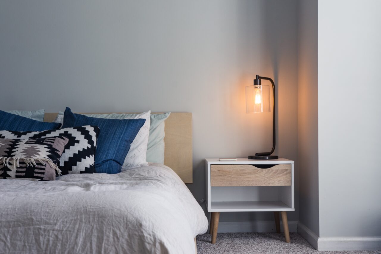 A nightstand with a light on next to a bed.